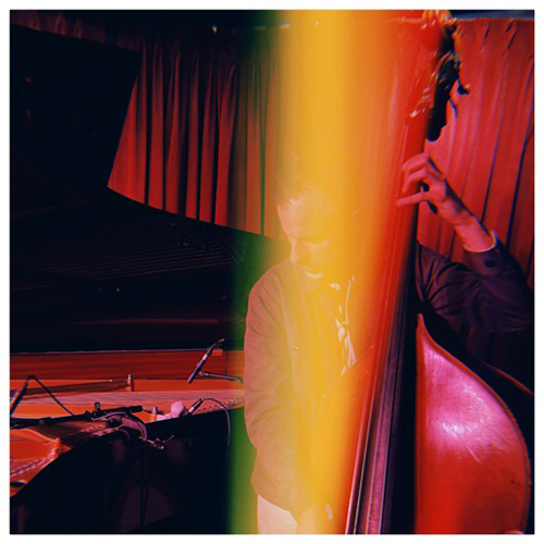 Warm red lighting fills the scene as a musician plays an upright bass beside a piano. A vertical streak of yellow and green light cuts through the image. Curtains in the background.