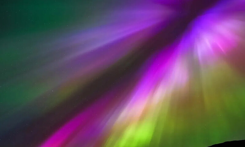 The cover of On The Edge (Of What Could Be) by Dave Bainbridge features a lone silhouette standing on a ridge beneath an aurora. Vivid streaks of purple, green, and pink light radiate across the night sky, converging in a burst of color. The artist’s name appears in ornate green lettering at the top left, while the album title rests at the bottom right in modern type.