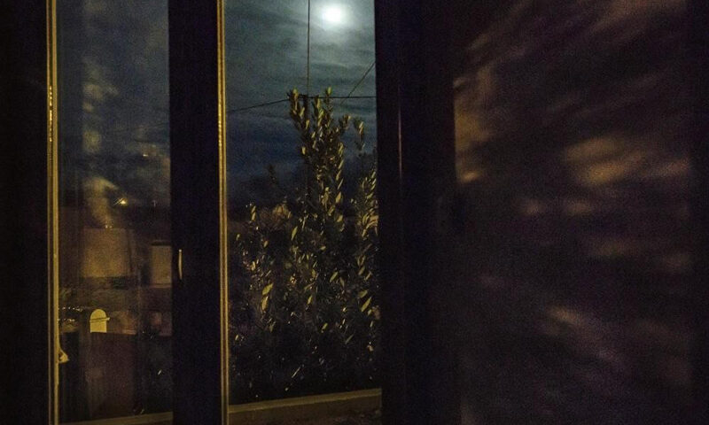 The cover of The Visitors by Gary Burton and Kirill Gerstein, featuring the music of Chick Corea. A darkened room frames a window through which a moonlit night is visible. The full moon, partially veiled by clouds, casts a subtle glow over a leafy tree just outside. Shadows and reflections play across the walls and glass.