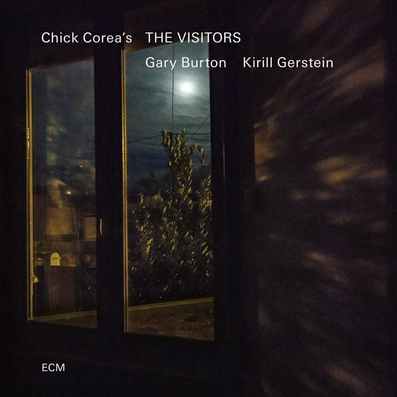 The cover of The Visitors by Gary Burton and Kirill Gerstein, featuring the music of Chick Corea. A darkened room frames a window through which a moonlit night is visible. The full moon, partially veiled by clouds, casts a subtle glow over a leafy tree just outside. Shadows and reflections play across the walls and glass.