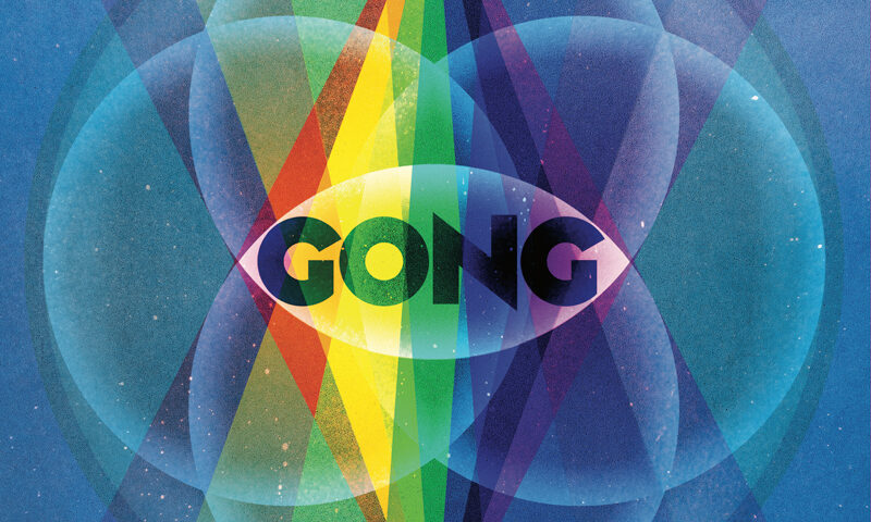 The cover of Bright Spirit by Gong presents a symmetrical, geometric design set against a deep blue background. At the center, a tall prism splits into bright rainbow beams that extend vertically through the composition. Overlapping translucent circles form a mandala-like pattern around the prism, creating an eye motif with the word “GONG” placed across its center. The album title appears below in spaced white letters.