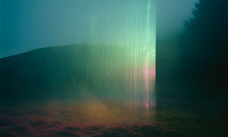 Johan Agebjorn - Southern-Forest cover artwork. A misty landscape fills the frame, tinted in deep teal and magenta hues. In the center, a tall glowing rectangular prism emits rainbow light, casting faint reflections onto the grassy ground. A dark hill rises in the background while a silhouette of trees stands at the right edge, partially swallowed by fog.