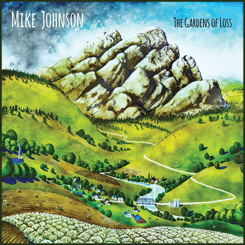 A pastoral painting dominates the cover, with rolling green hills, winding pale roads, and a cluster of tiny homes gathered near a small bridge and river. Towering craggy mountains rise in the background beneath a clear sky. The artist’s name, Mike Johnson, sits in the upper left, while the album title, The Gardens of Loss, appears in the upper right.