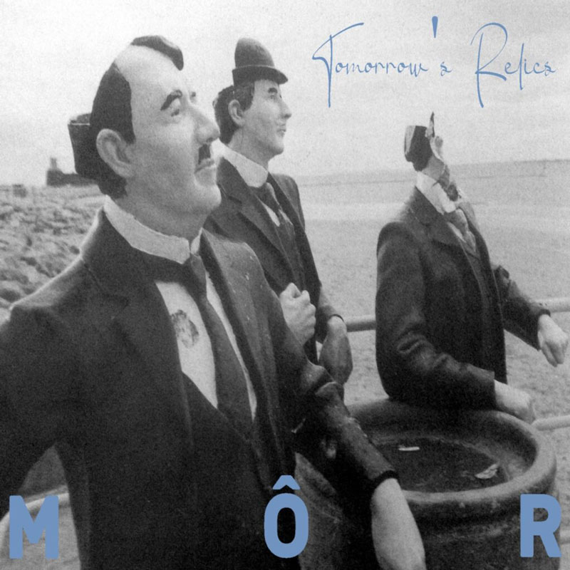 A grainy black-and-white seaside photograph shows three suited figures with mannequin-like heads, posed mid-stride along a coastal railing. Handwritten blue script reading Tomorrow’s Relics floats in the upper right, while the band name appears spaced across the bottom in pale blue capitals.