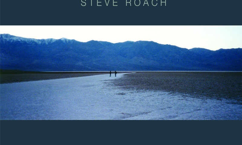 The album cover for New Life Dreaming (2024 Remaster) by Steve Roach features a landscape photograph of a vast, desolate plain with two distant figures walking along a narrow water path. A mountain range looms in the background under a pale sky.