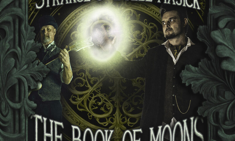 The cover of Strange & Wylde Majick by The Book of Moons features two men in vintage attire, one holding a guitar and the other conjuring a glowing orb of light. Ornate green foliage frames the scene, with an intricate golden circular design in the background. The title appears in bold, gothic-style lettering beneath the artists’ names.