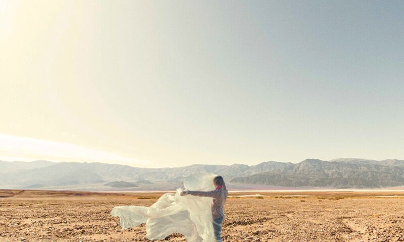 Time’s Forgotten – “Songs of Awakening” cover atwork. a photo of a woman standing in a flat desertlike zone holding flowing white cloth.