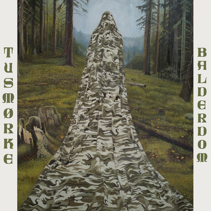 A cloaked figure draped in camouflage cloth stands in a misty forest clearing, the long fabric pooling across the mossy ground. Tall trees and cut stumps surround the figure. The band name “TUSMØRKE” runs down the left side and the title BALDERDOM down the right in green lettering.