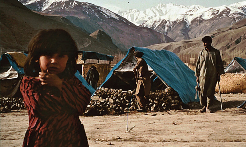 The album cover for Volker Lankow’s 14 Years Through A Turmoiled World features a young girl in the foreground, while men and makeshift shelters with blue tarps occupy the background, set against snow-capped mountains.